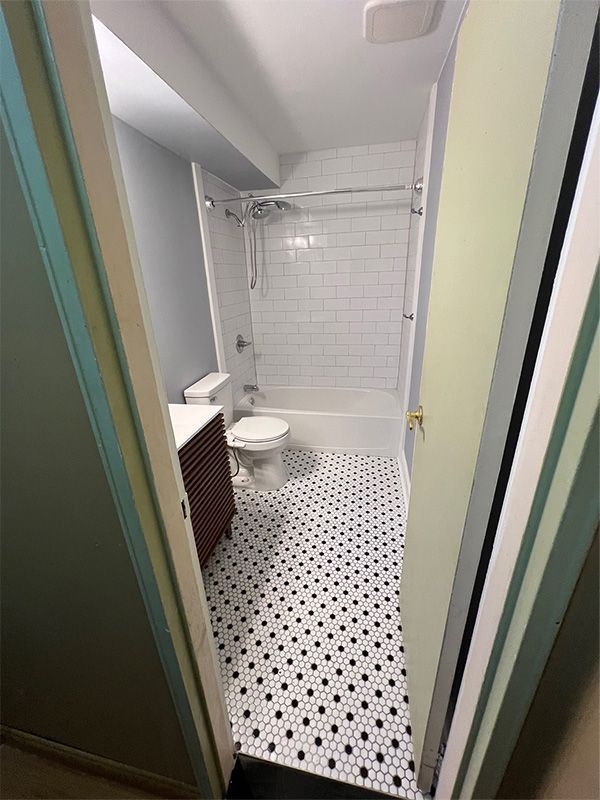 Narrow bathroom with white tile walls, black-and-white floor, shower, toilet, and sink visible through the doorway