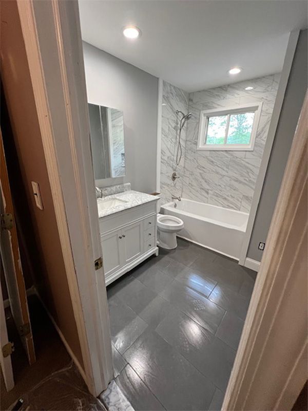 Small gray bathroom with vanity, toilet, bathtub, marble-style shower wall, and a window above the tub.