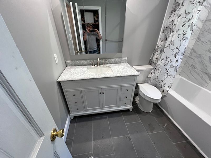 Small bathroom with vanity, marble countertop, mirror, gray tile floor, and bathtub with floral shower curtain.