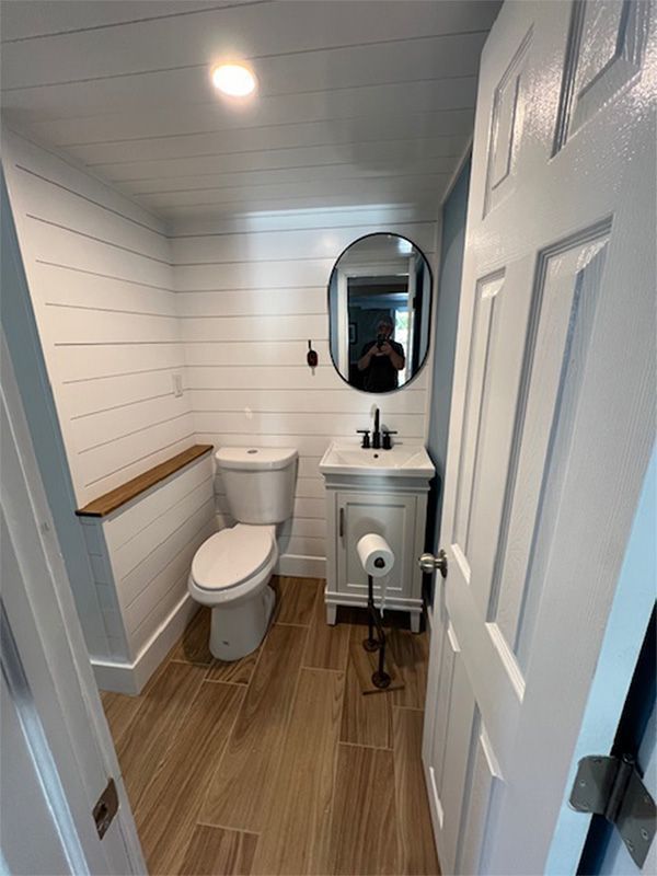 Narrow white bathroom with toilet, pedestal sink, oval mirror, and wood-look floor.