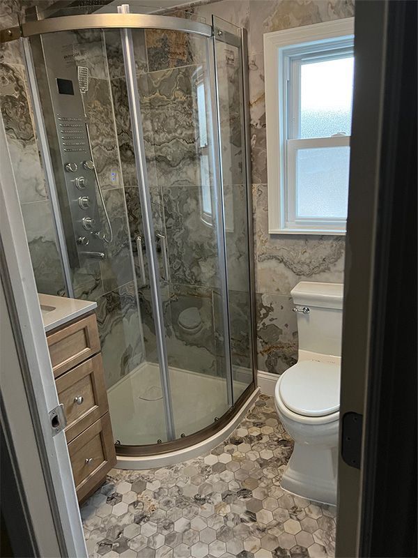 Small bathroom with curved glass shower, mosaic tile floor, vanity, and toilet beside a window.