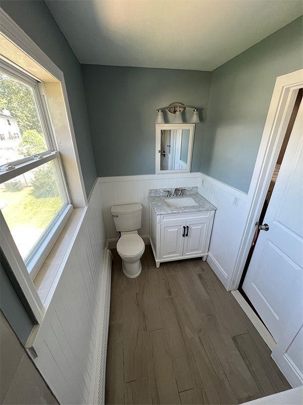 Narrow bathroom with white toilet and vanity, gray walls, wood-look floor, and window on the left.
