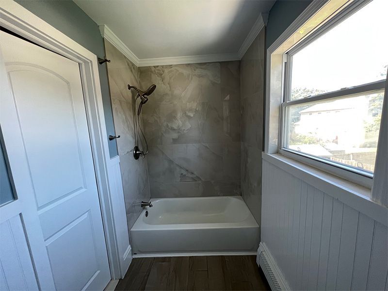 Small bathroom with a white tub, gray tiled shower wall, window, and white paneled door.