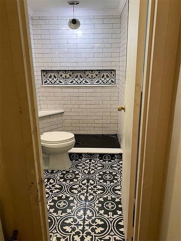 Narrow view into a tiled bathroom with a toilet, patterned black-and-white floor, and white subway walls.