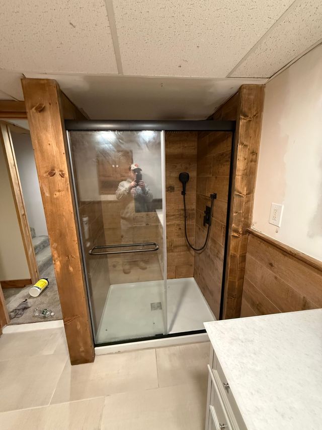 Glass shower stall in a rustic bathroom with wood paneling and a white vanity.