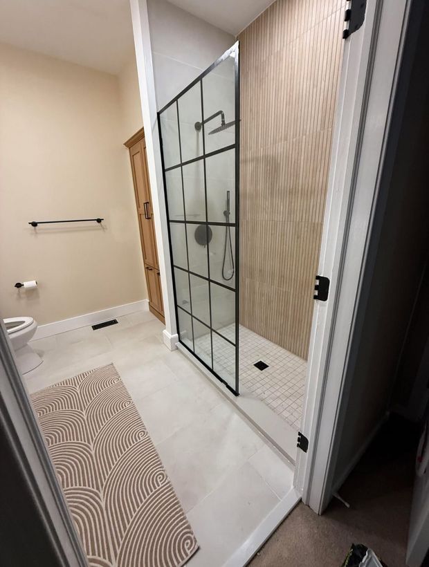 Modern bathroom with glass shower, beige tile, towel rack, and patterned bath mat.