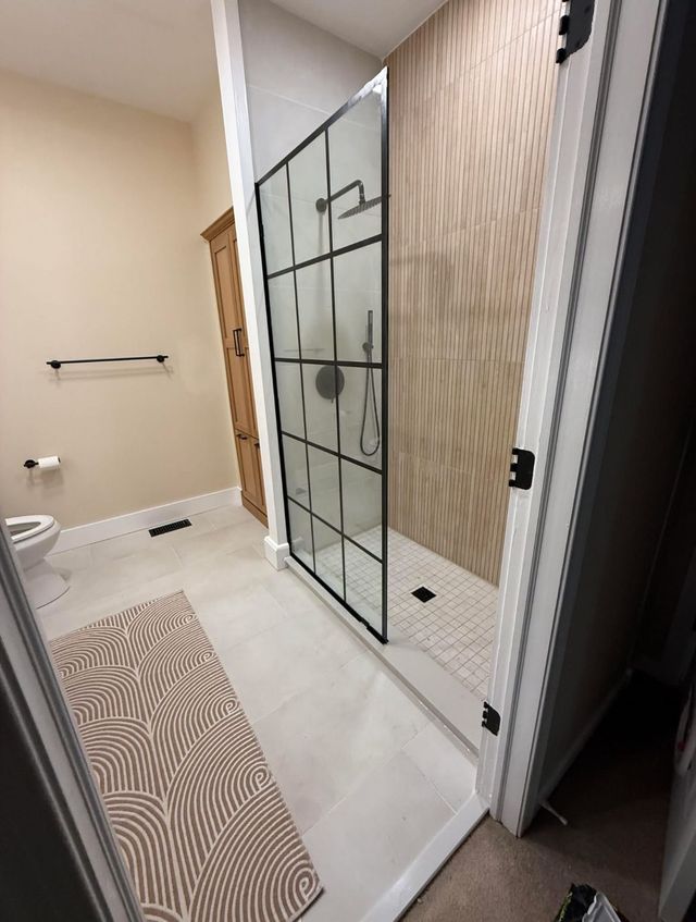 Modern bathroom with glass shower, beige tile, towel rack, and patterned bath mat.