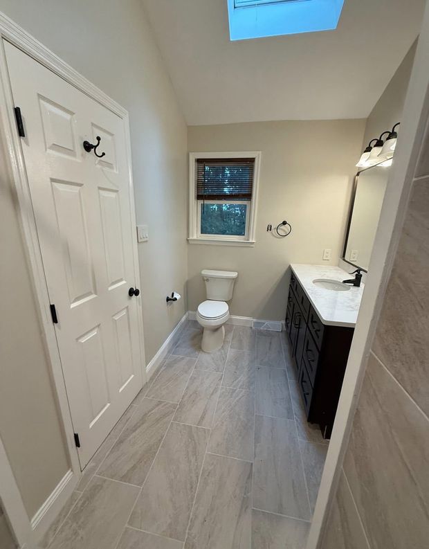 Narrow bathroom with white walls, skylight, toilet, and dark vanity with sink.