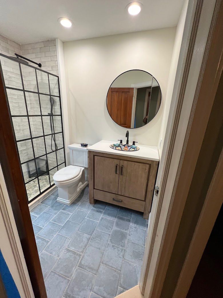 Small bathroom with tiled floor, toilet, wood vanity, round mirror, shower curtain, and soft ceiling lights