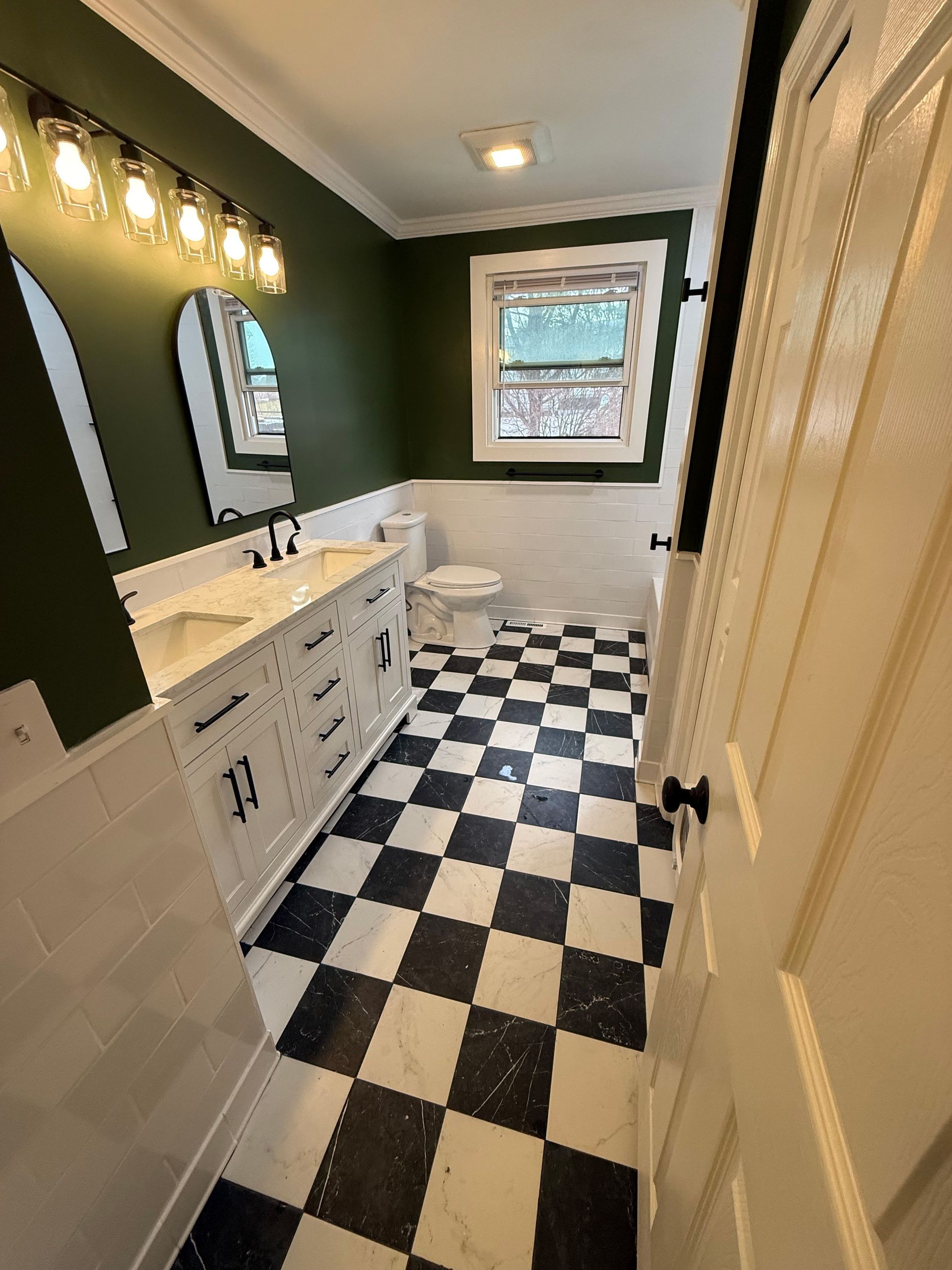 Small bathroom with black-and-white checkered floor, dark green walls, white vanity, and window light