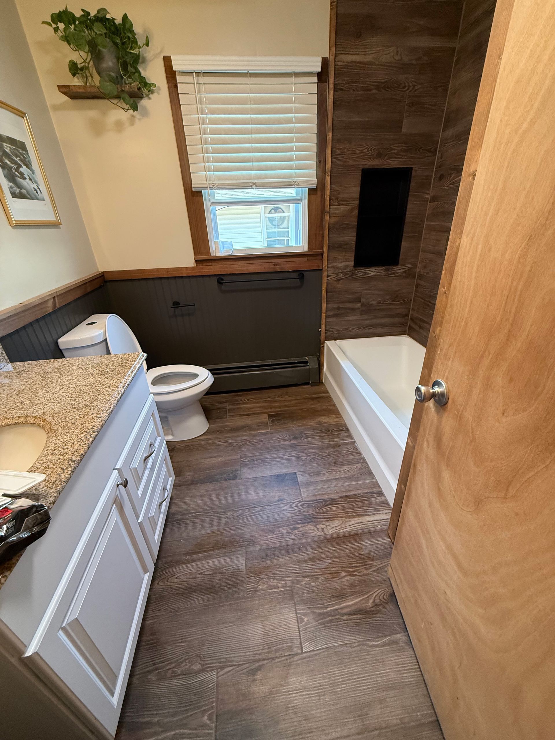 Small bathroom with wood vanity, toilet, bathtub, window, and dark tile accent wall.