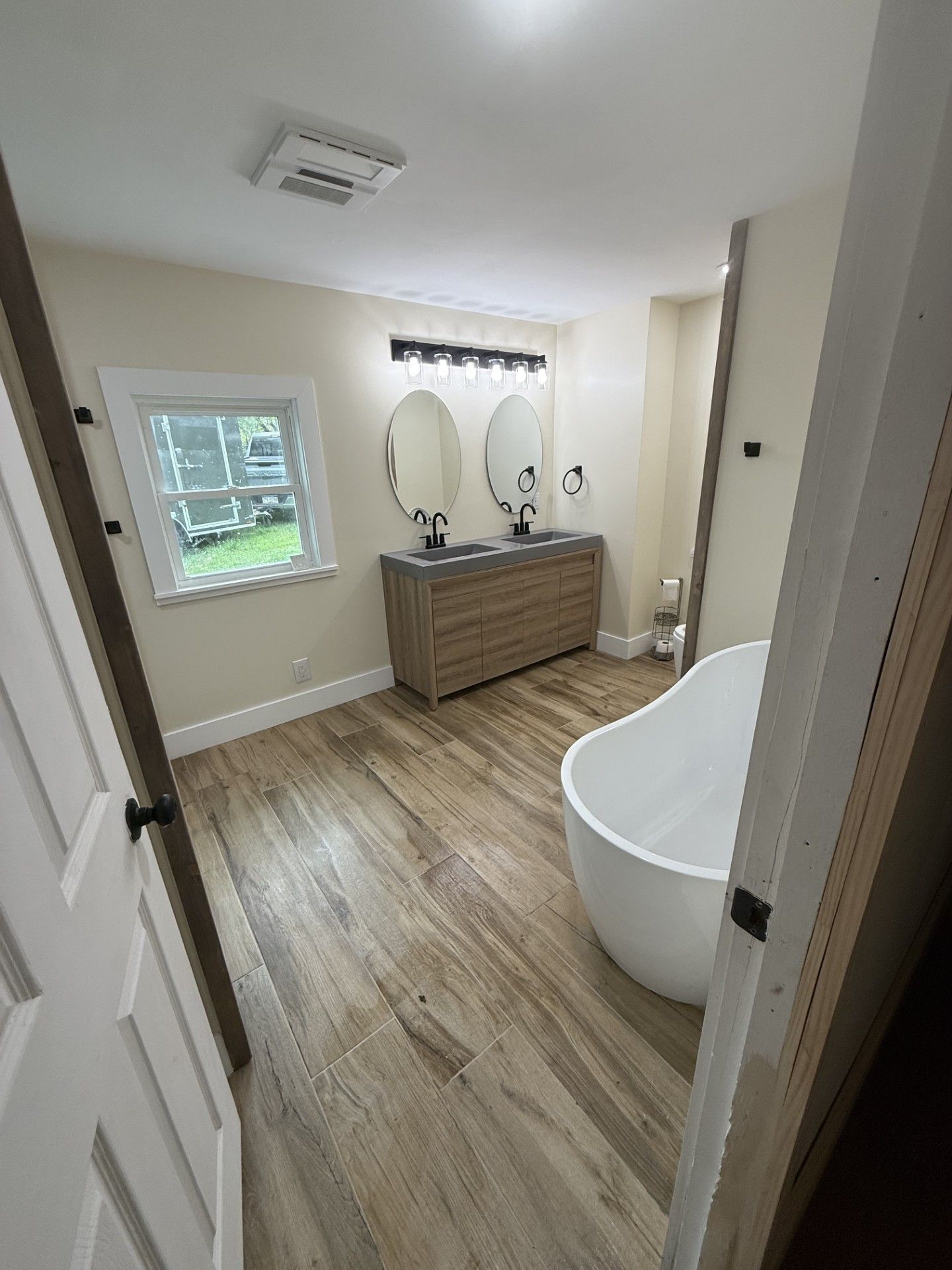 Bright bathroom with wood vanity, oval mirrors, white tub, and light wood-look floor.