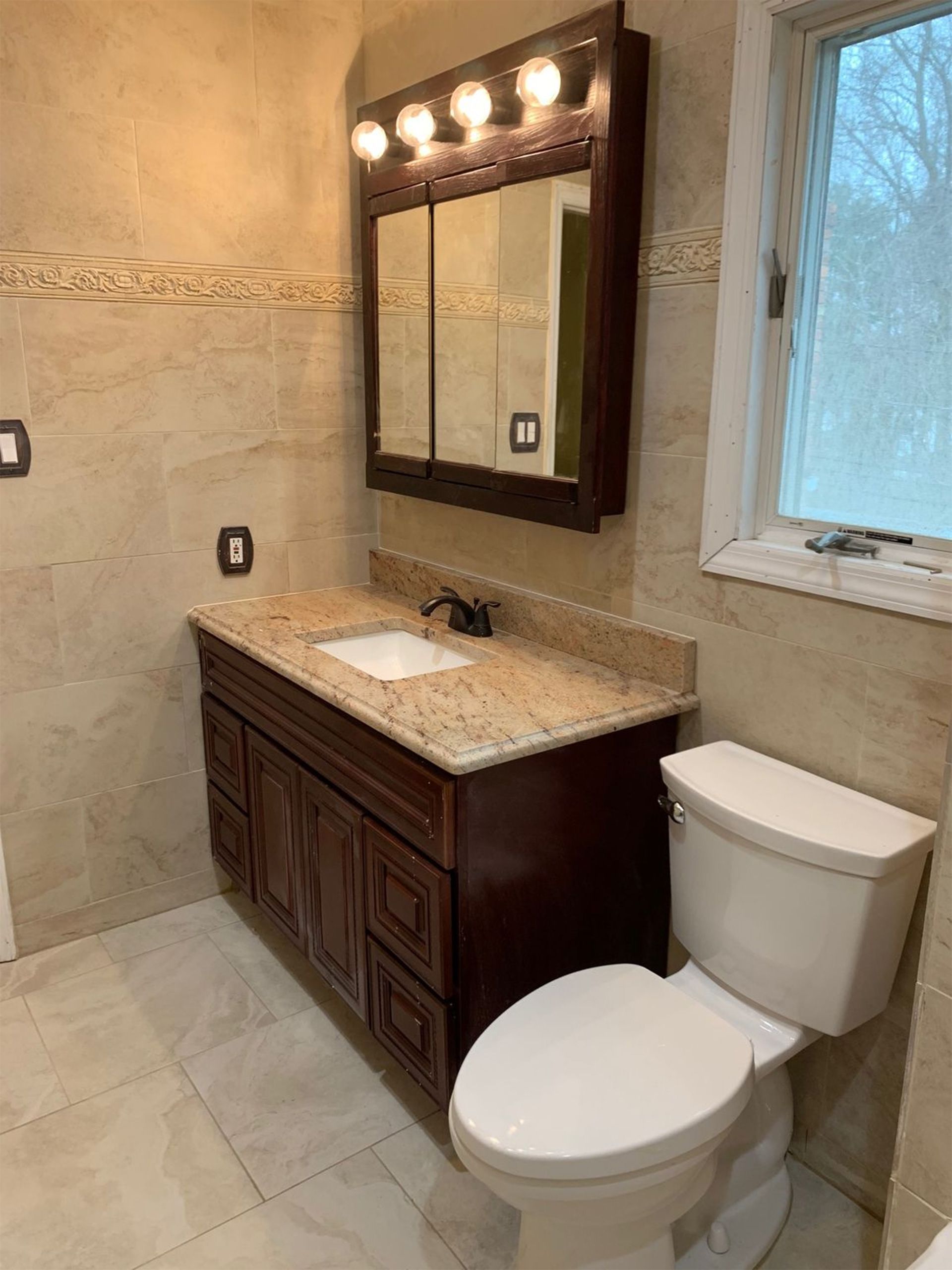 Bathroom with granite vanity, lit mirror, white toilet, and tiled walls beside a window.