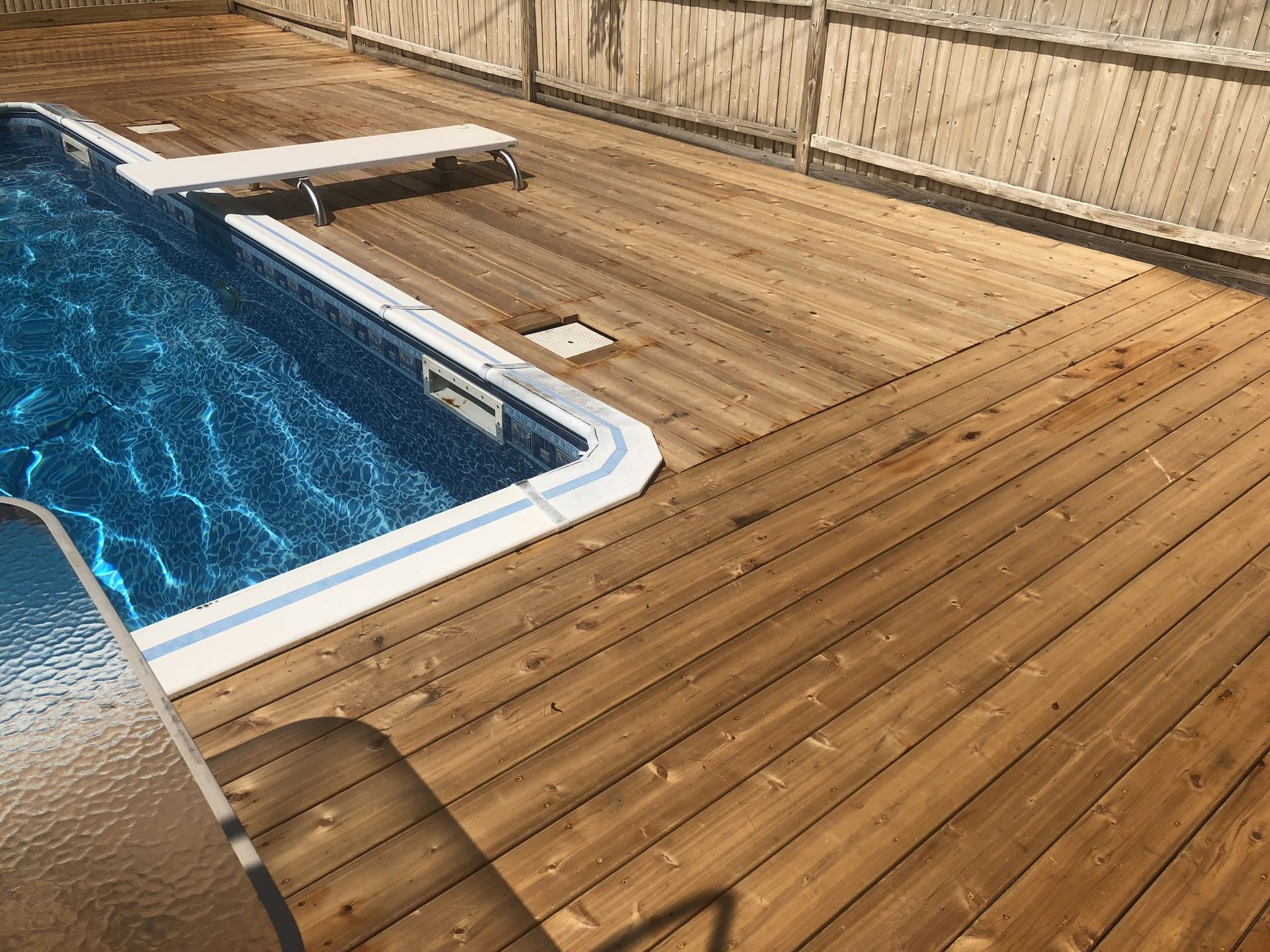 Wooden pool deck with blue swimming pool and a diving board by a fenced wall