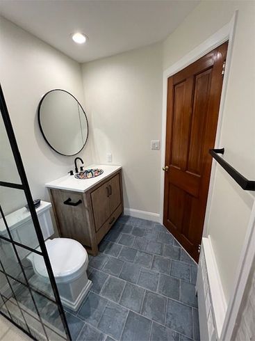 Small bathroom with gray tile, wood vanity, round mirror, toilet, and wooden door.