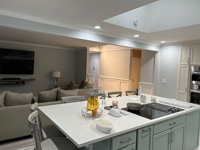 Open kitchen and living room with gray sofa, wall TV, and white island counter with fruit centerpiece.