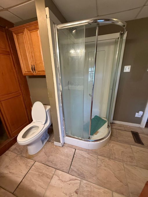 Bathroom with corner glass shower, white toilet, beige tile floor, and wood cabinets