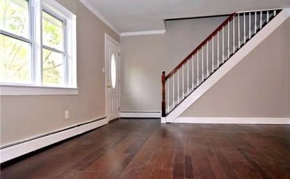 Empty living room with hardwood floors, white walls, and staircase by a front door and window.