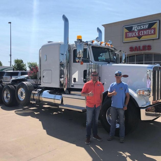 Two men standing in front of a silver semi truck at a truck center sales lot