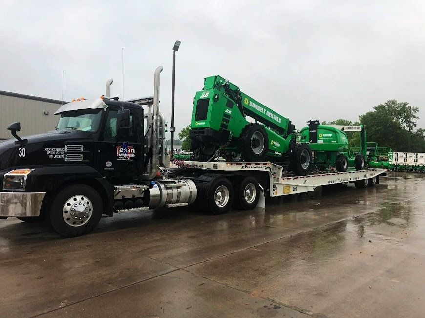 Black semi hauling green construction equipment on a flatbed trailer in a wet parking lot