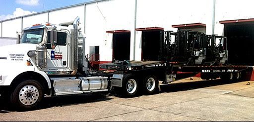 Flatbed tow truck carrying forklifts outside a warehouse row
