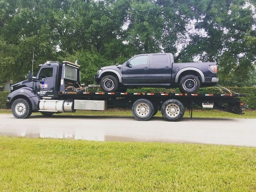 Black tow truck carrying a gray pickup on a flatbed beside a grassy roadside