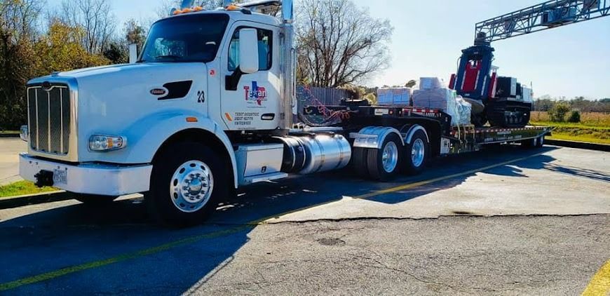 White semi-truck hauling a long trailer on a sunny roadside, with construction equipment in back