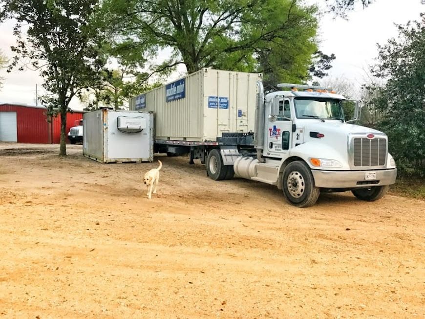 White semi-truck with trailer parked on a dirt lot beside a small white building and trees, with a dog nearby