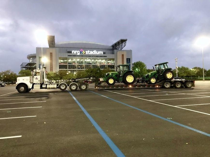 White truck hauling two green tractors on a flatbed in a parking lot at dusk