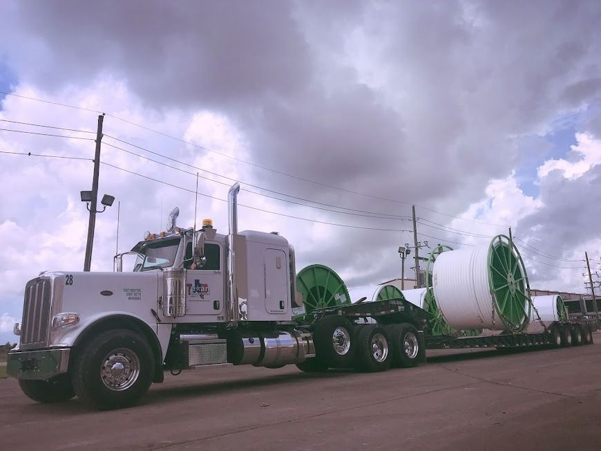 White semi-truck hauling a large industrial reel on a flatbed under a cloudy sky