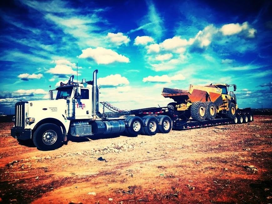 White semi-truck hauling a yellow excavator on a flatbed trailer across a dirt road under a bright blue sky