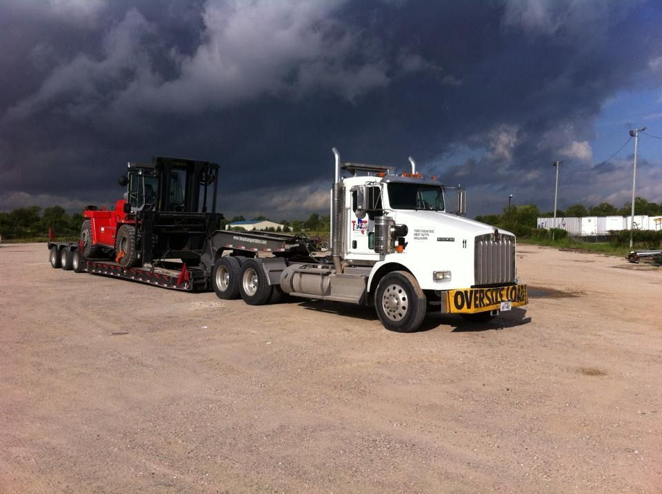White semi-truck hauling a black construction machine on a trailer under a dark stormy sky