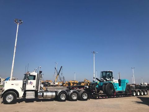 White semi truck hauling a teal excavator on a lowboy trailer in an industrial yard