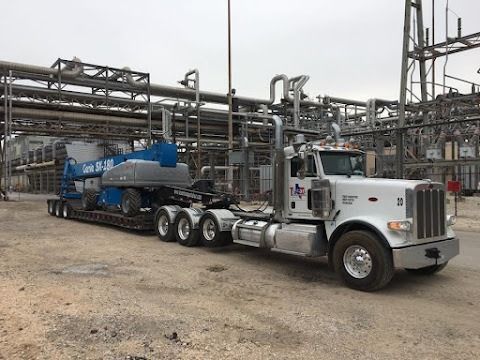 White semi-truck hauling a blue tanker trailer at an industrial plant, with pipes and structures in the background