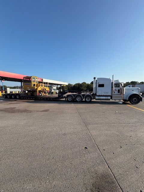 White semi truck with flatbed trailer parked at a gas station under a clear blue sky.