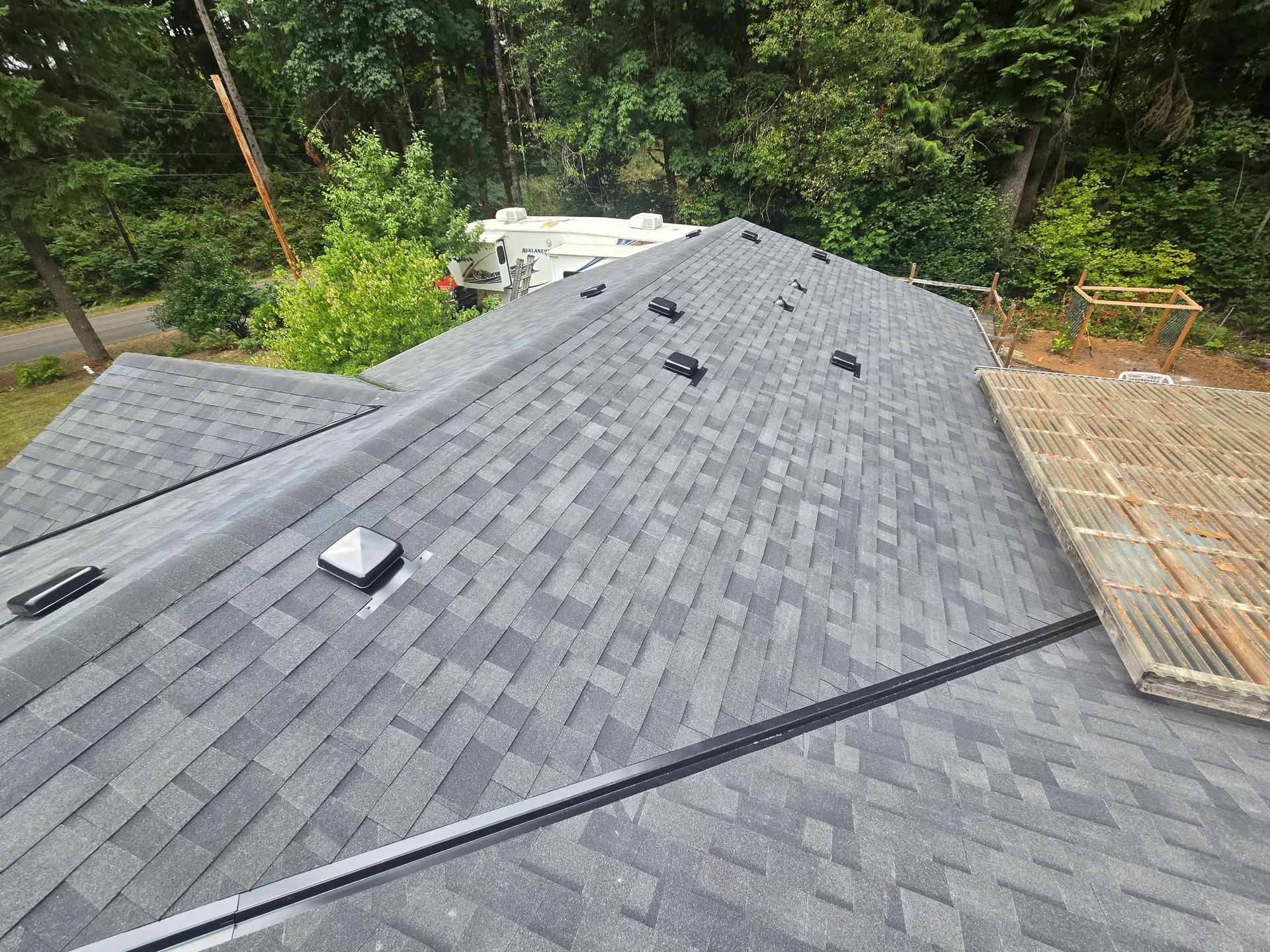 Gray shingled roof under construction with vents, skylights, and trees in the background