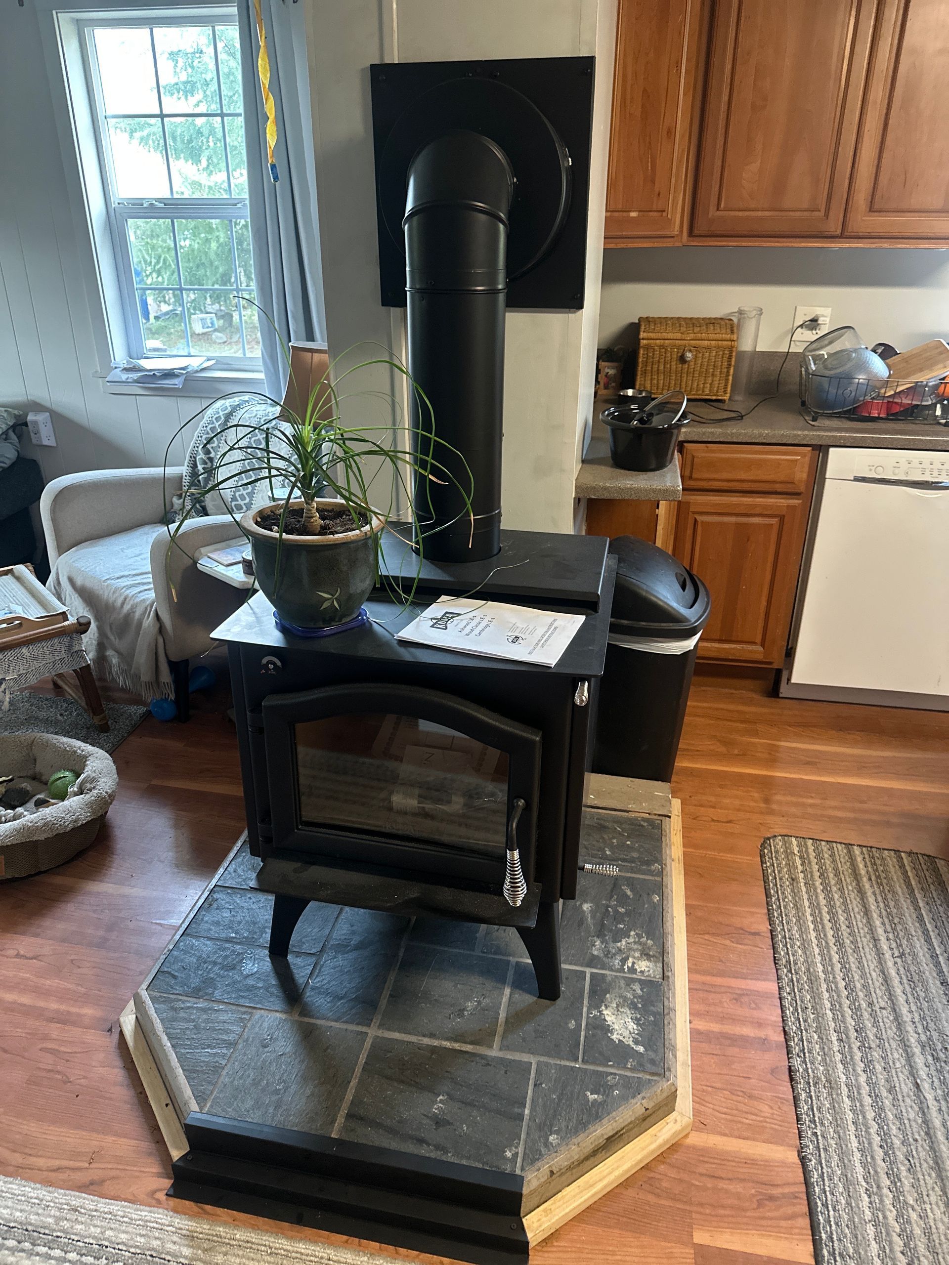 A wood-burning stove in a kitchen setting with black stovepipe, tile hearth, and surrounding furniture.
