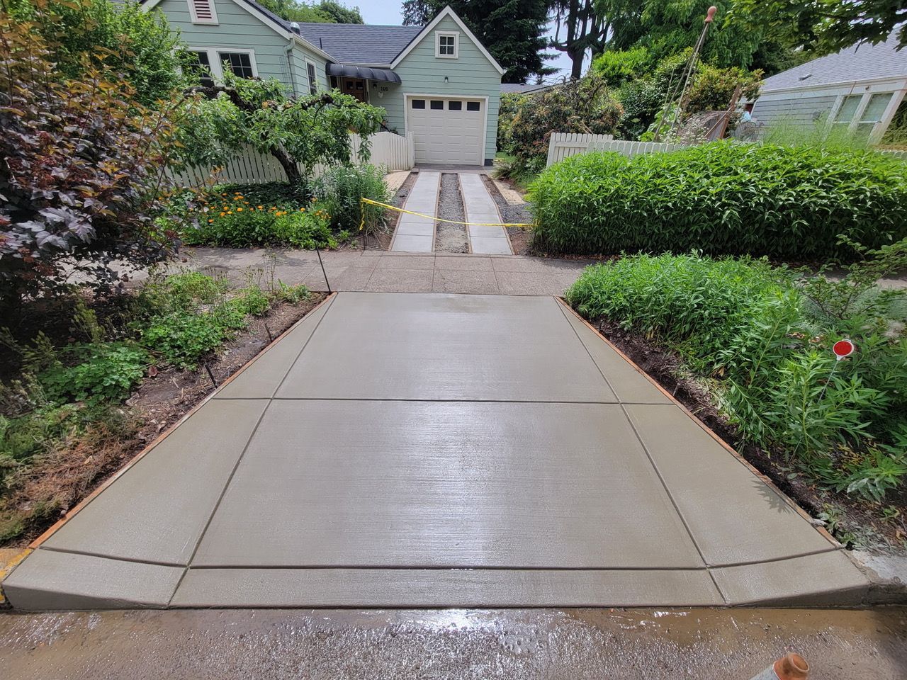 Wet concrete driveway leading to a garage, bordered by green shrubs and garden beds.