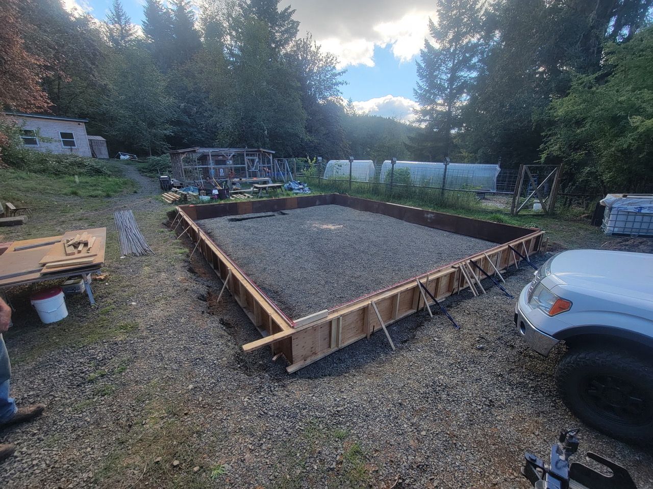 Fresh gravel foundation with wooden forms in a yard, surrounded by trees and parked vehicles