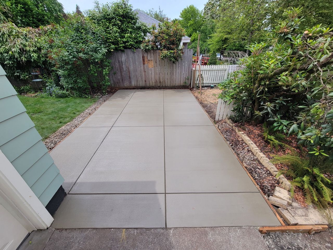 Concrete backyard patio path leading to a wooden gate, bordered by shrubs and garden plants
