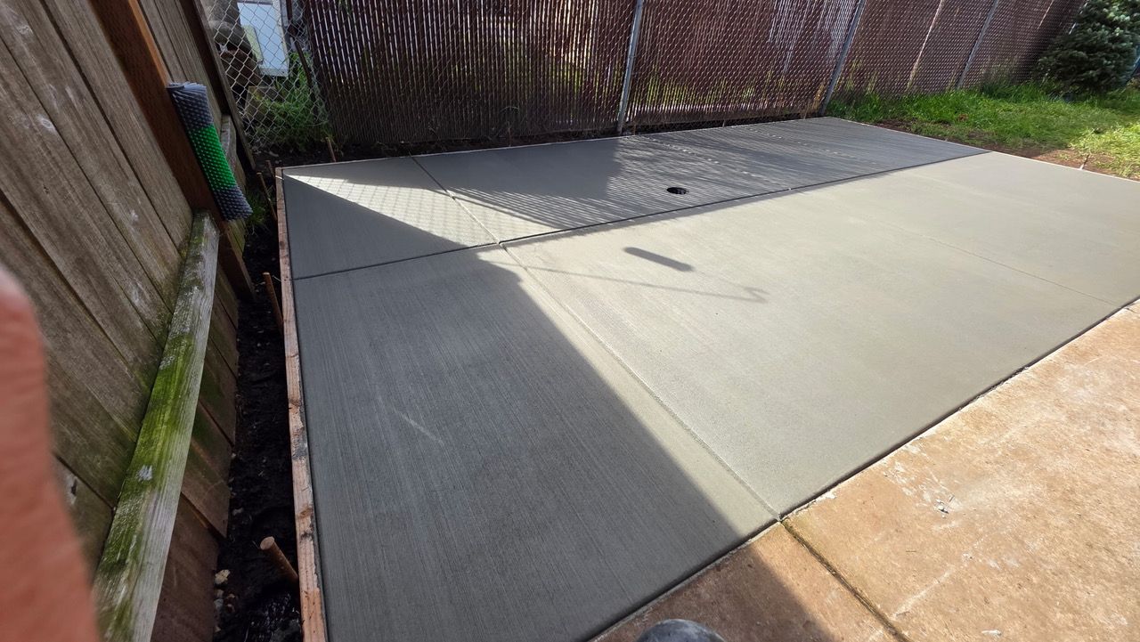 Concrete patio beside a wooden fence, with a small drain and sunlight across the surface.