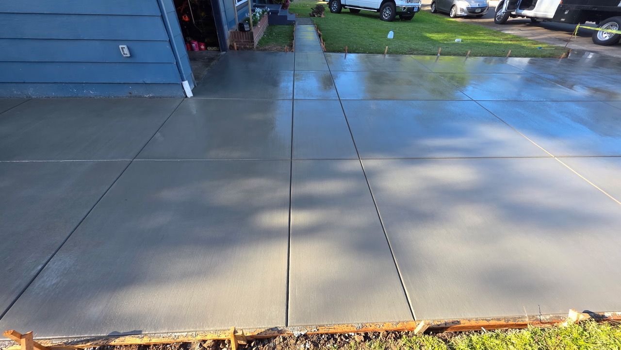 Concrete driveway and sidewalk beside a blue house, with tree shadows and a wet patch near the top.