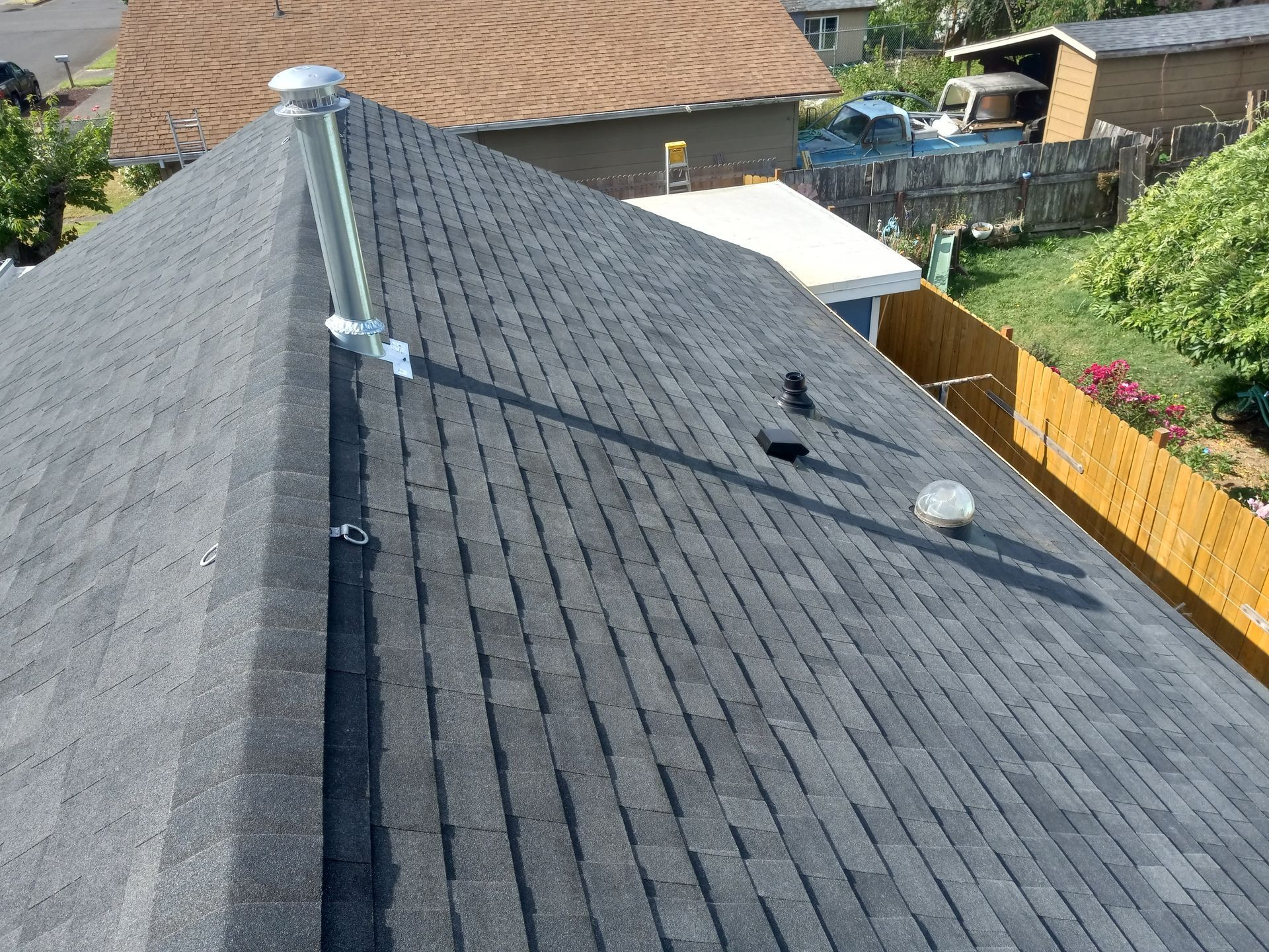 Gray shingled roof with a chimney and vents, viewed from above beside a fenced backyard.