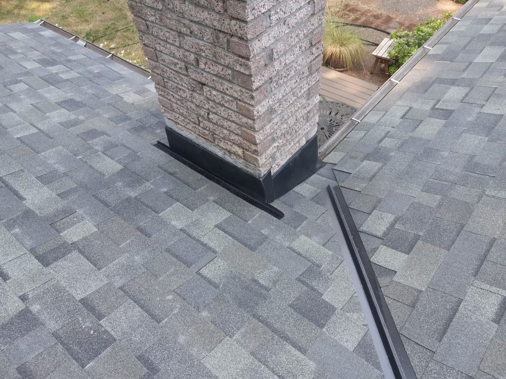 Gray roof shingles around a brick chimney with black flashing and roof seams