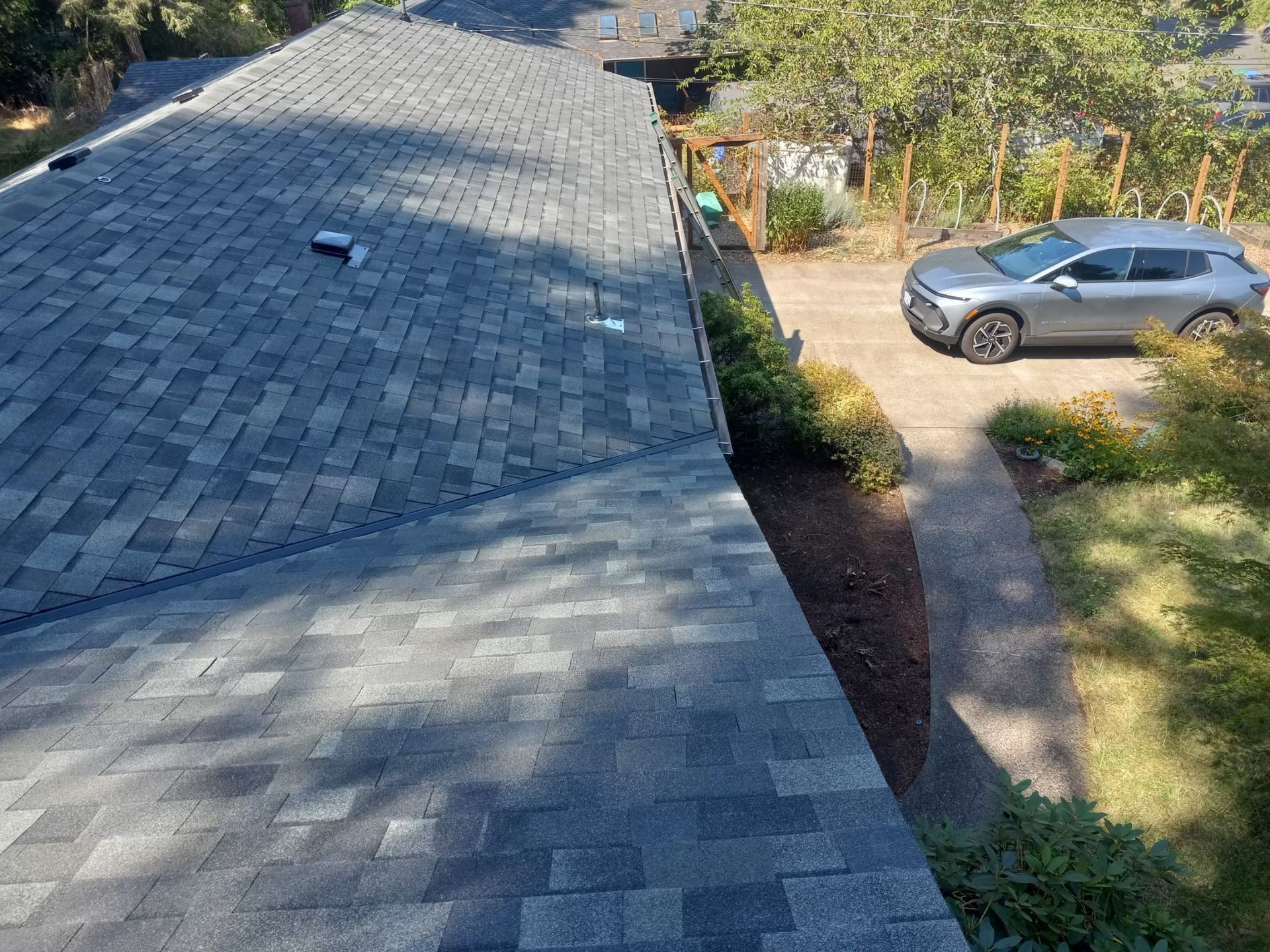Roof shingles on a house with a driveway, parked silver car, and trees in the yard.