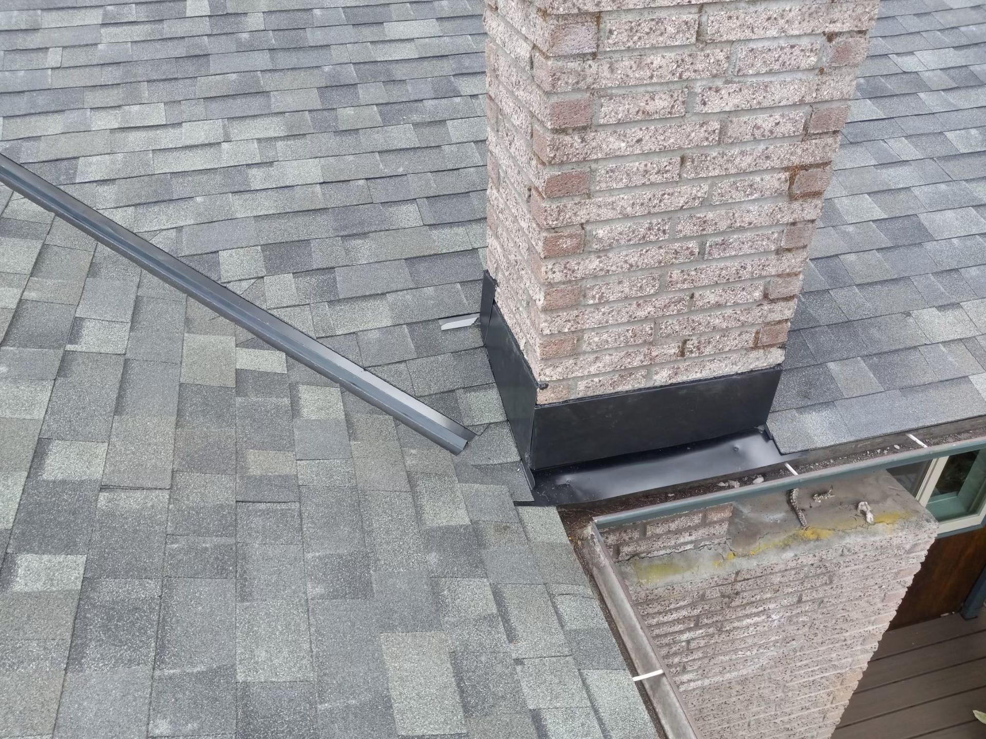 Rooftop shingles beside a brick chimney and metal flashing around a roof opening