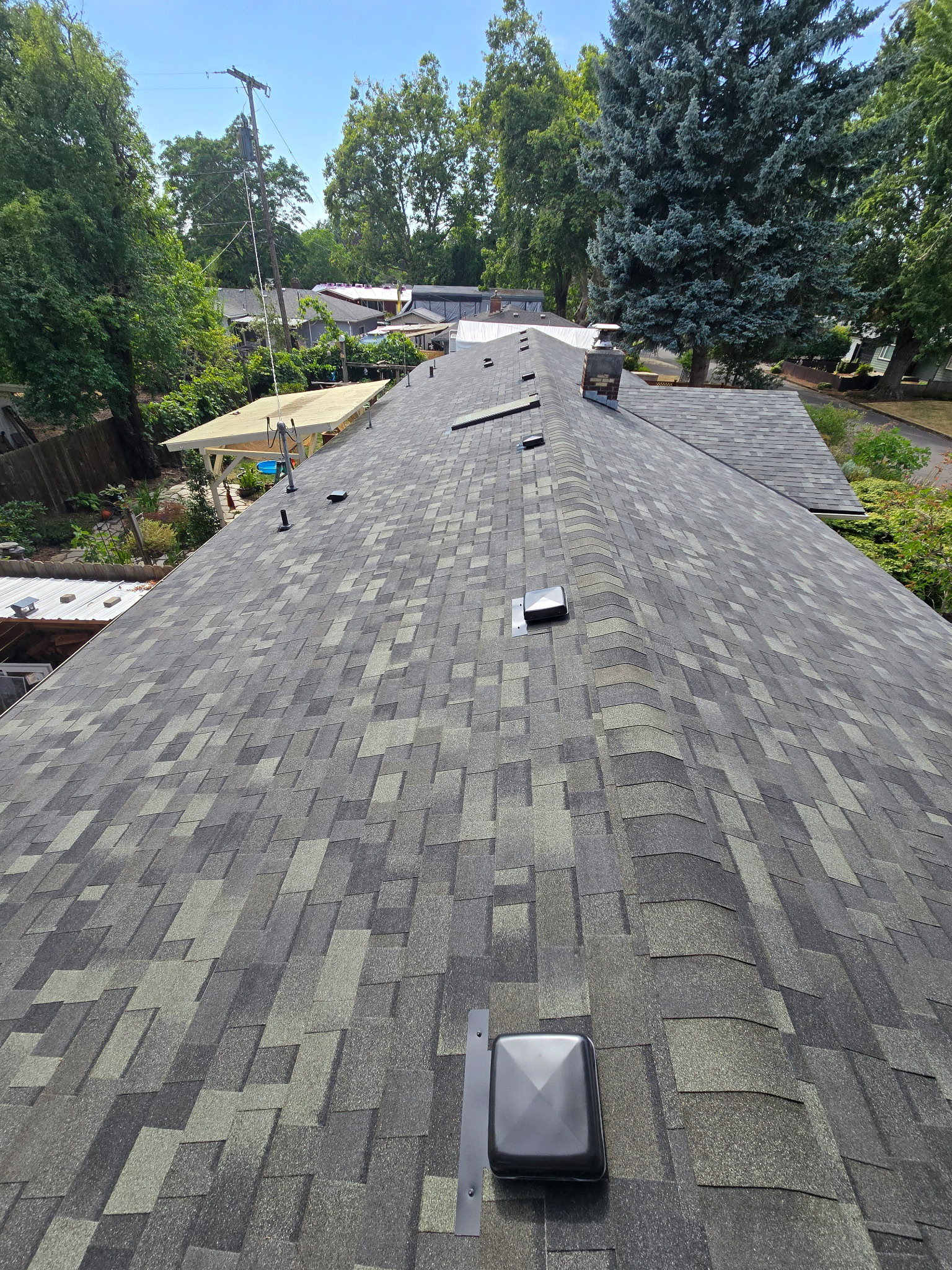 Rooftop with gray shingles and vents, viewed from above, with trees and neighboring houses nearby