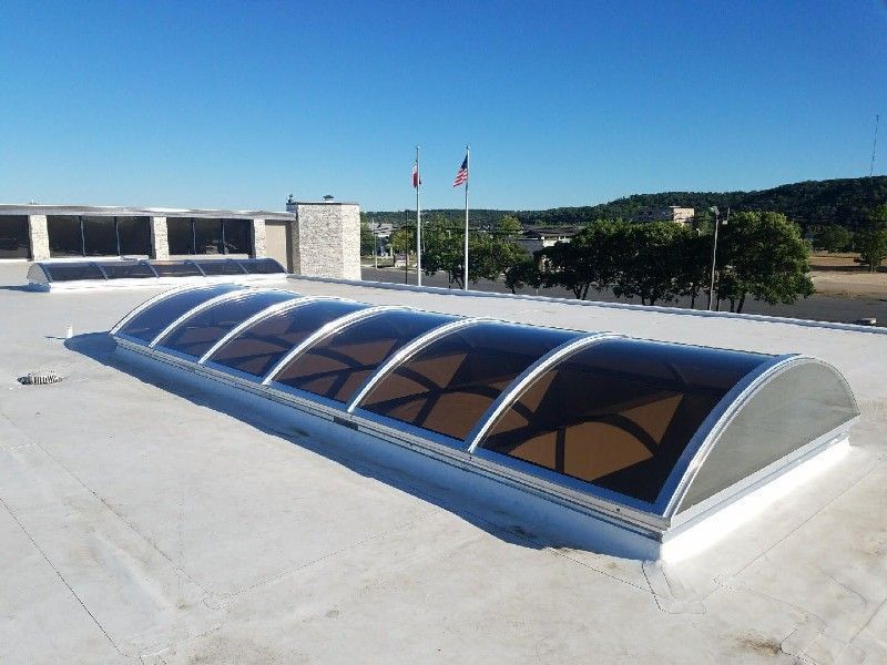 Skylight on a flat roof, arched brown panels, with buildings and flags in the background on a sunny day.