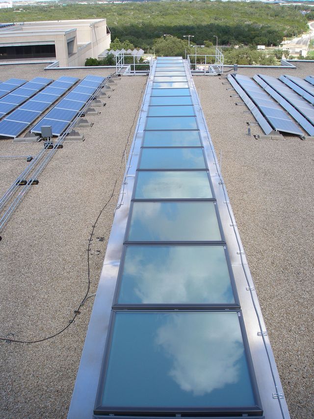 Skylight on a rooftop, framed by metal, reflecting the sky. Solar panels nearby.