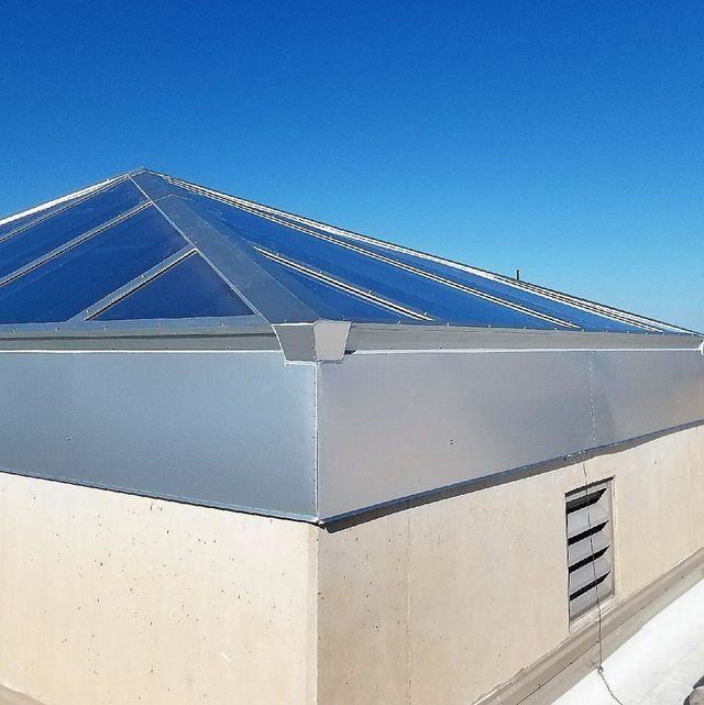 A skylight on a building roof with a clear blue sky background. The skylight has a metallic frame and blue-tinted panels.
